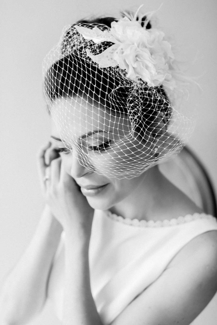 Black-and-white portrait of a bride looking down with a soft smile, one hand at her cheek, wearing a birdcage veil with a large fabric flower.