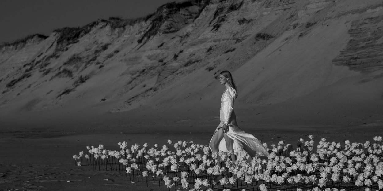 Black-and-white fashion-style editorial of a woman in a flowing white dress walking through a field of white roses at the foot of a sand dune.