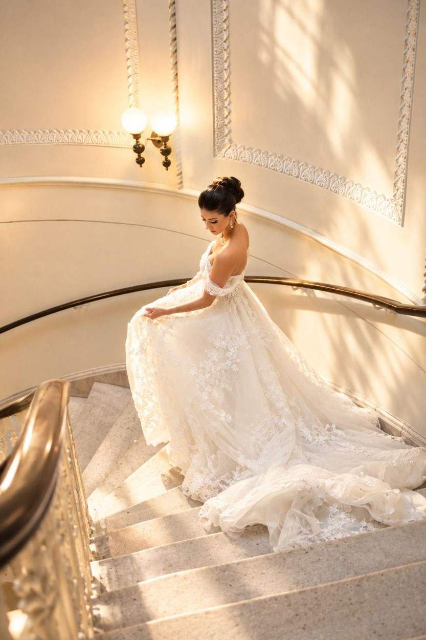 A bride in an off-the-shoulder lace gown gathers her skirt as she descends a curved marble staircase, her long train spilling over the steps in warm window light.