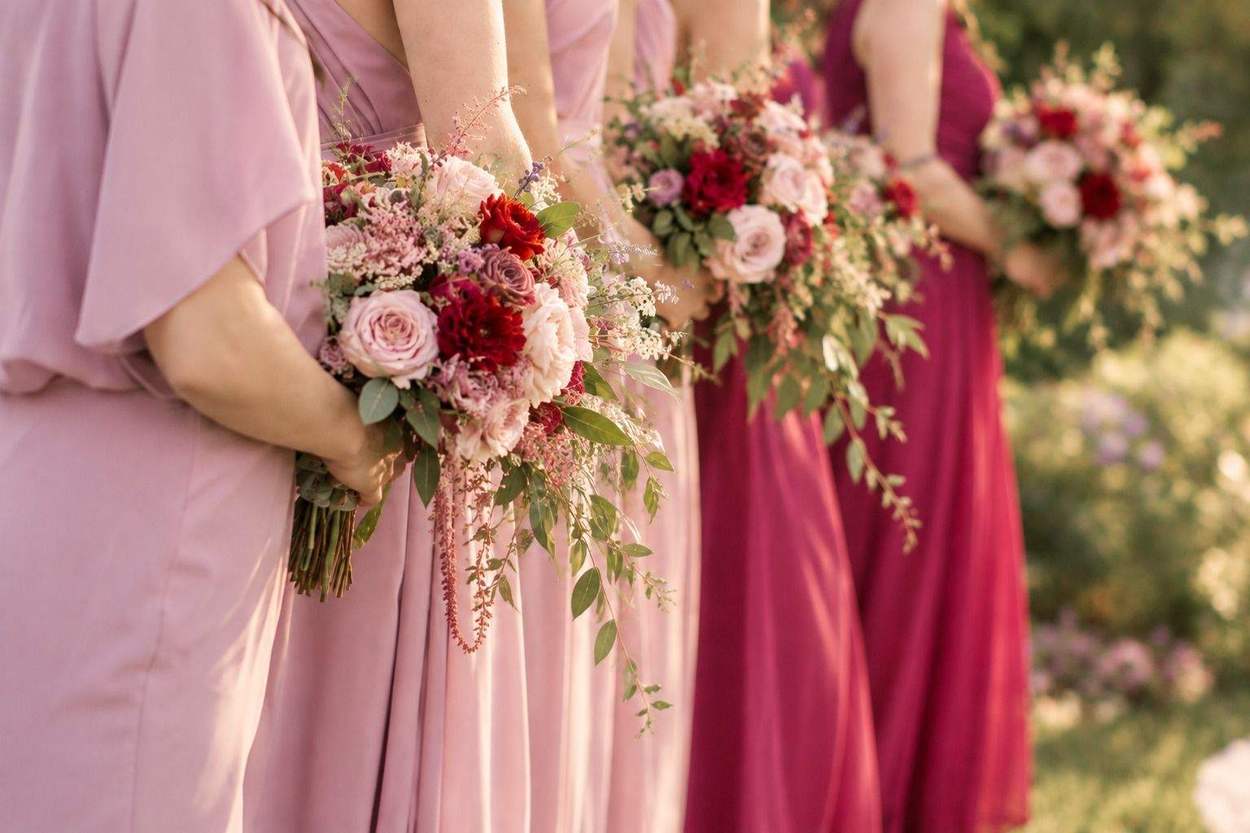 Bridesmaids in shades of dusty rose to raspberry hold trailing bouquets of pink and red roses and dahlias behind their backs.