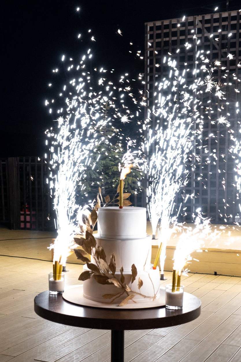 A three-tier white wedding cake decorated with gold leaves, surrounded by tall white sparkler fountains at night on a wooden terrace.