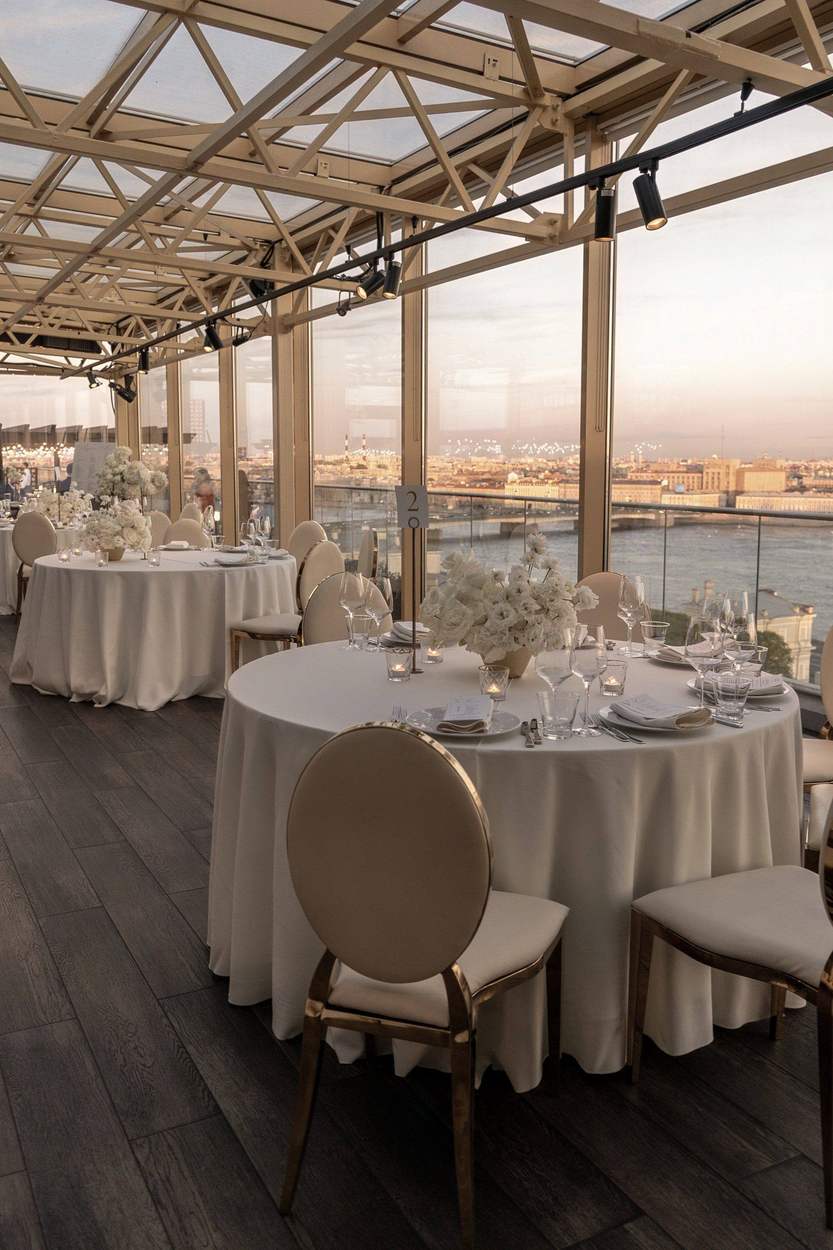 Round reception tables dressed in ivory linens with white floral centerpieces and candlelight, set beneath a glass-and-steel roof with a river and city skyline glowing at sunset.