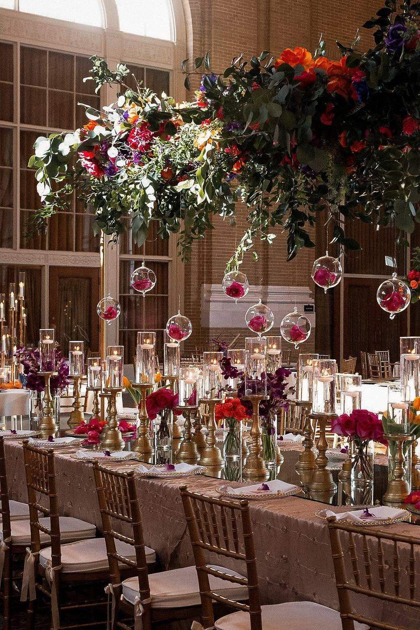 A long reception table in a brick hall with hanging glass globes holding roses, gold candlesticks, floating candles and a canopy of orange, magenta and purple flowers.