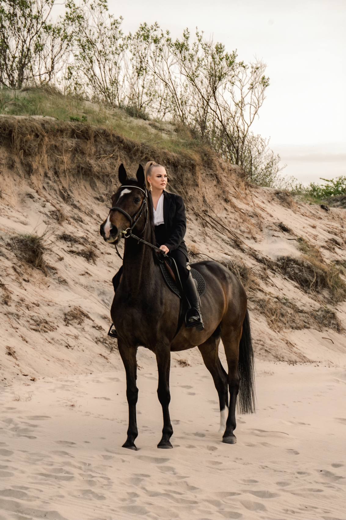 Woman in a black blazer sitting on a dark horse on a sandy slope with dune grasses behind her