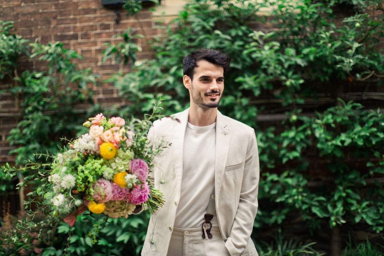 Smiling man in a cream linen jacket carrying a large bouquet of peonies and ranunculus in front of a brick wall covered in green vines.