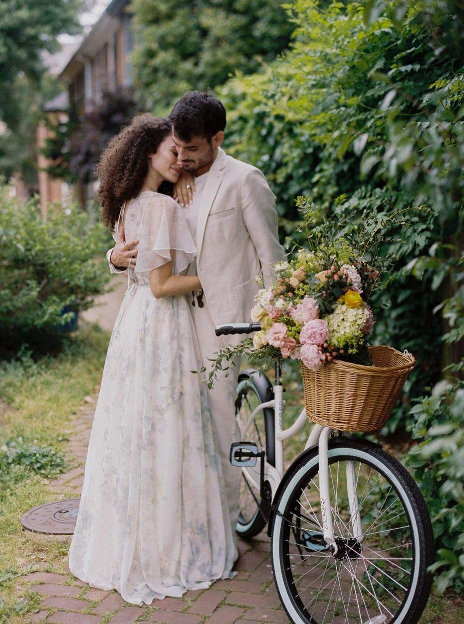 Couple embracing on a brick path beside a white bicycle with a wicker basket of pink and yellow flowers and a leafy hedge behind them.