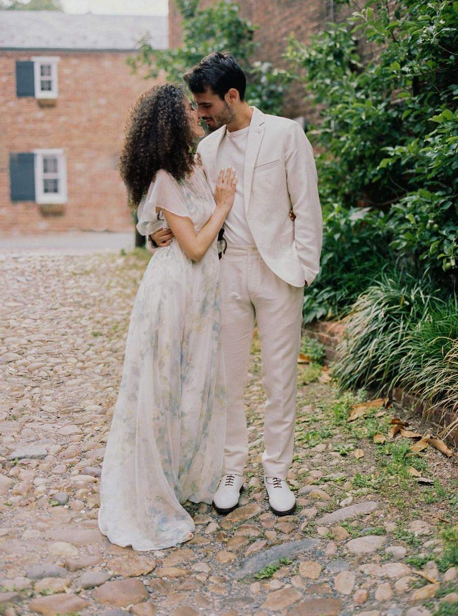 Full-length portrait of a couple about to kiss on a cobblestone street, she in a floral chiffon gown and he in a cream suit and white shoes, a brick townhouse behind.