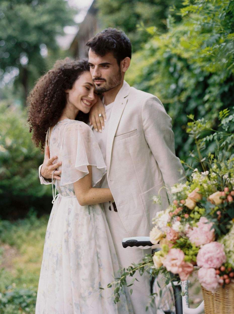 Close embrace between a smiling woman in a floral chiffon dress and a man in a cream jacket, a basket of pink peonies in the foreground and green hedges behind.