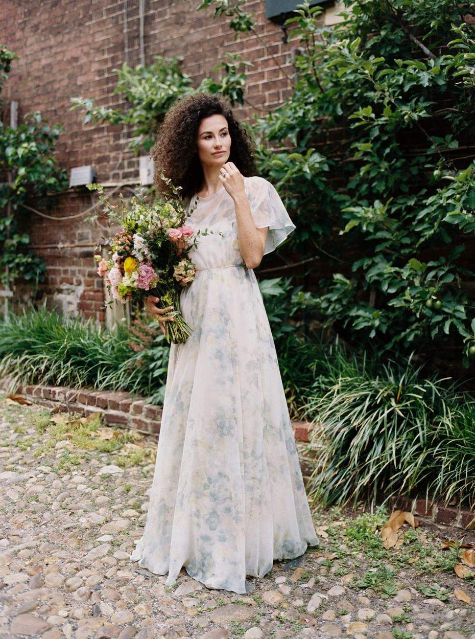 Woman in a pale blue floral chiffon dress holding a loose bouquet of pink peonies, ranunculus and eucalyptus on a cobblestone lane beside a brick wall.