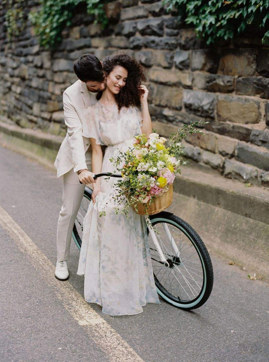 Man in a cream linen suit leaning in to kiss his fiancée's neck as she sits on a bicycle with a basket of peonies beside an ivy-topped stone wall.