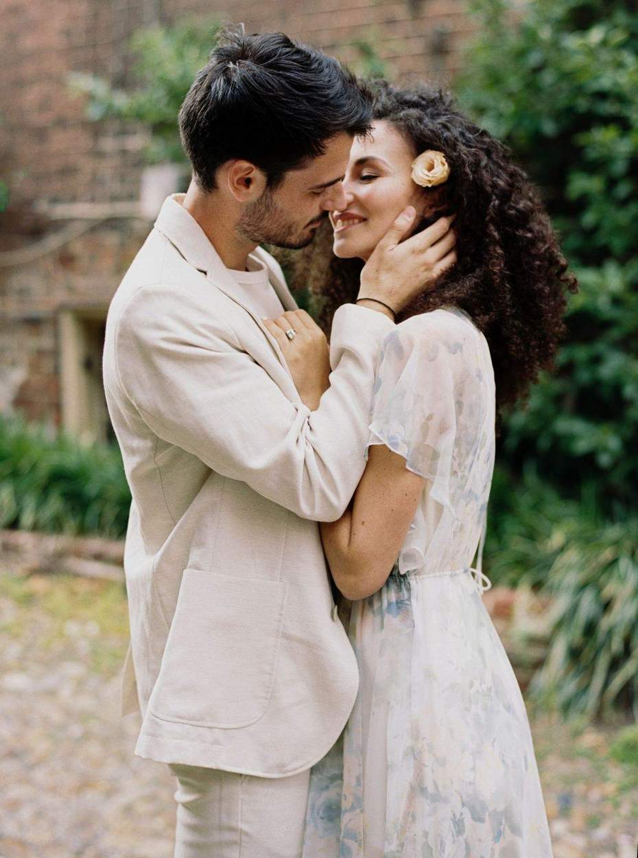 Couple nose to nose just before a kiss, she with a peach lisianthus in her curls and he in a cream linen jacket, soft green garden behind them.