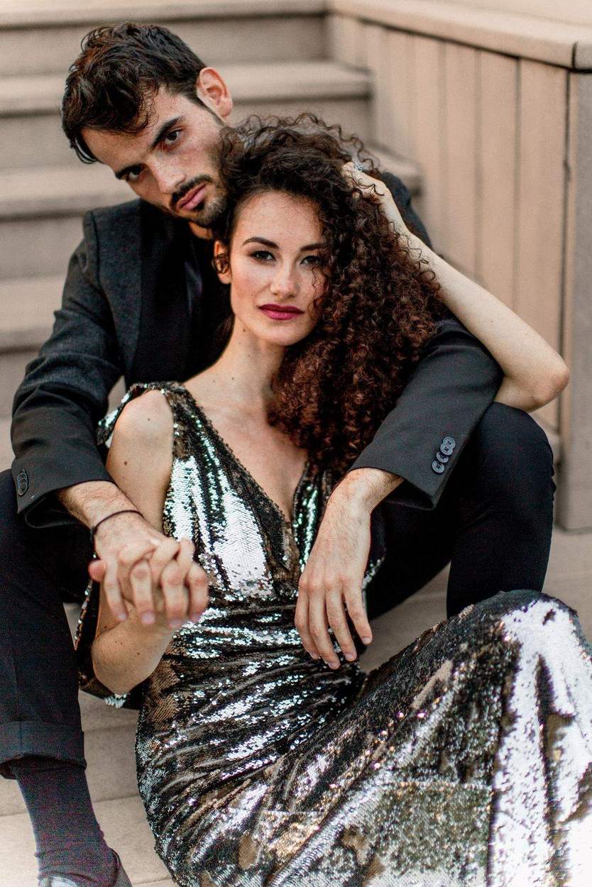 Engaged couple seated close on stone steps holding hands, he in a black suit and she in a sequin gown, Alexandria VA.