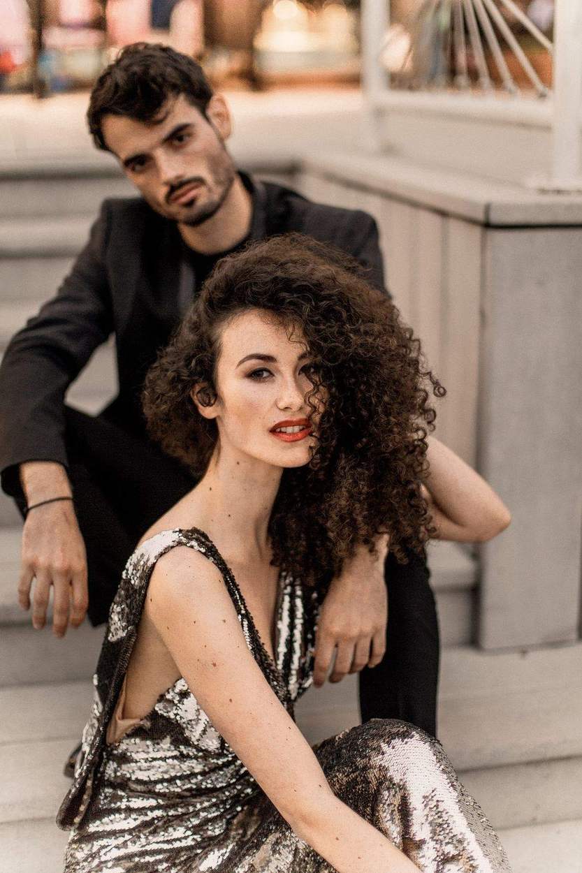 Man in a black jacket seated behind a woman in a sequin gown on stone steps, both looking into the camera, Old Town Alexandria engagement photos.