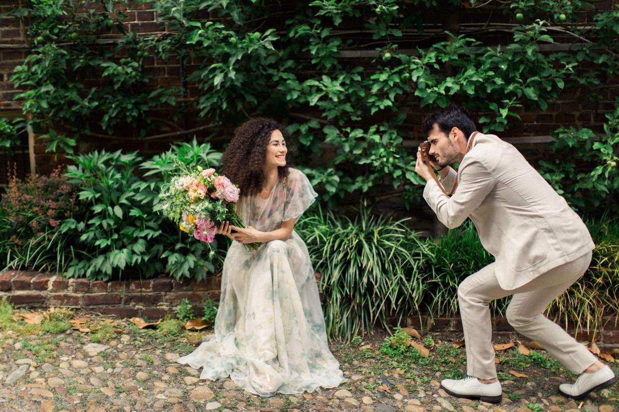 Woman laughing with a loose bouquet as her fiancé crouches to photograph her with a film camera in front of a brick wall and green foliage.