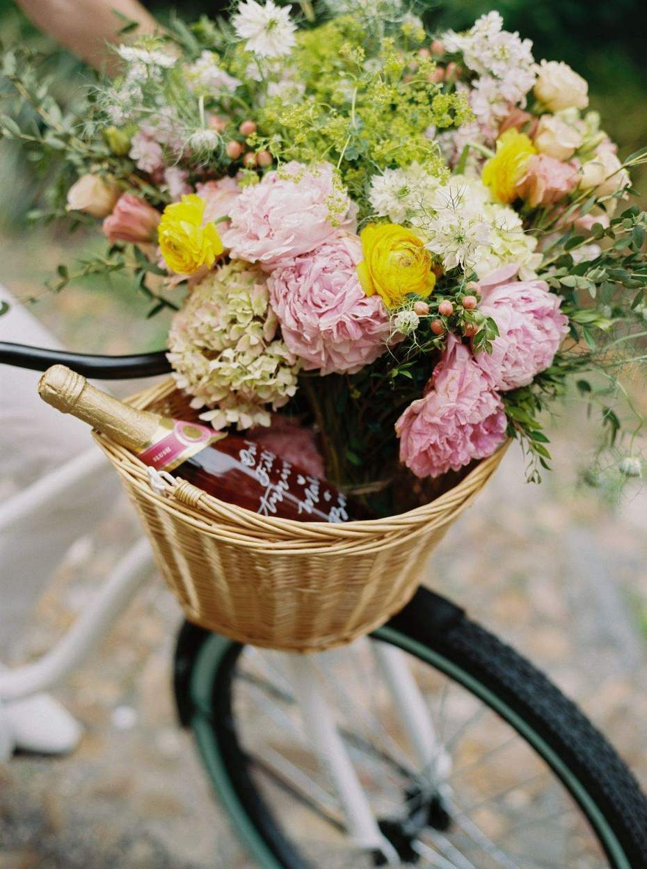 Bottle of blush sparkling wine with a hand-lettered label tucked into a bicycle basket of peonies, hydrangea and yellow ranunculus.