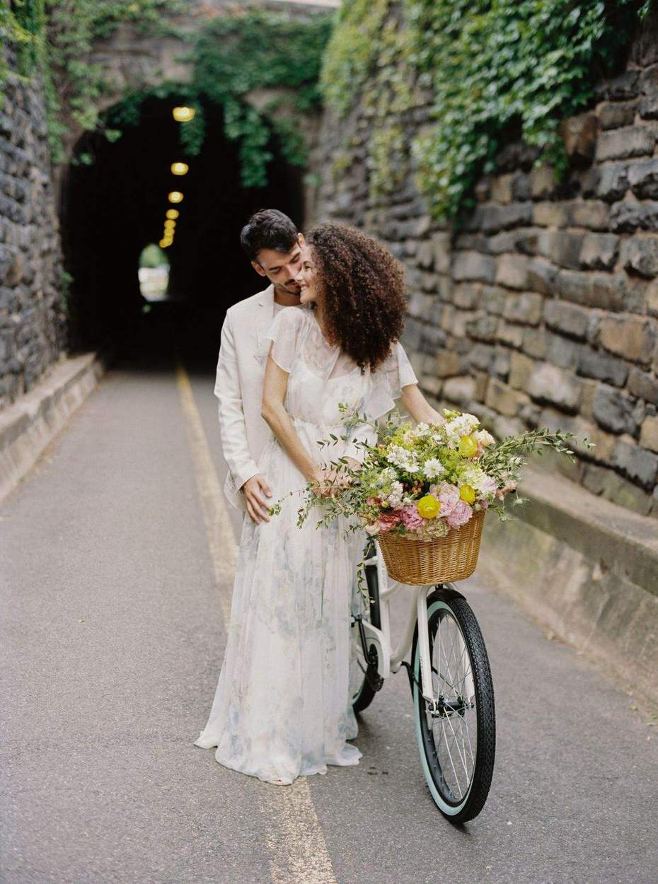 Engaged couple embracing beside a white bicycle with a flower-filled basket on a paved path in front of an ivy-covered stone tunnel in Old Town Alexandria.
