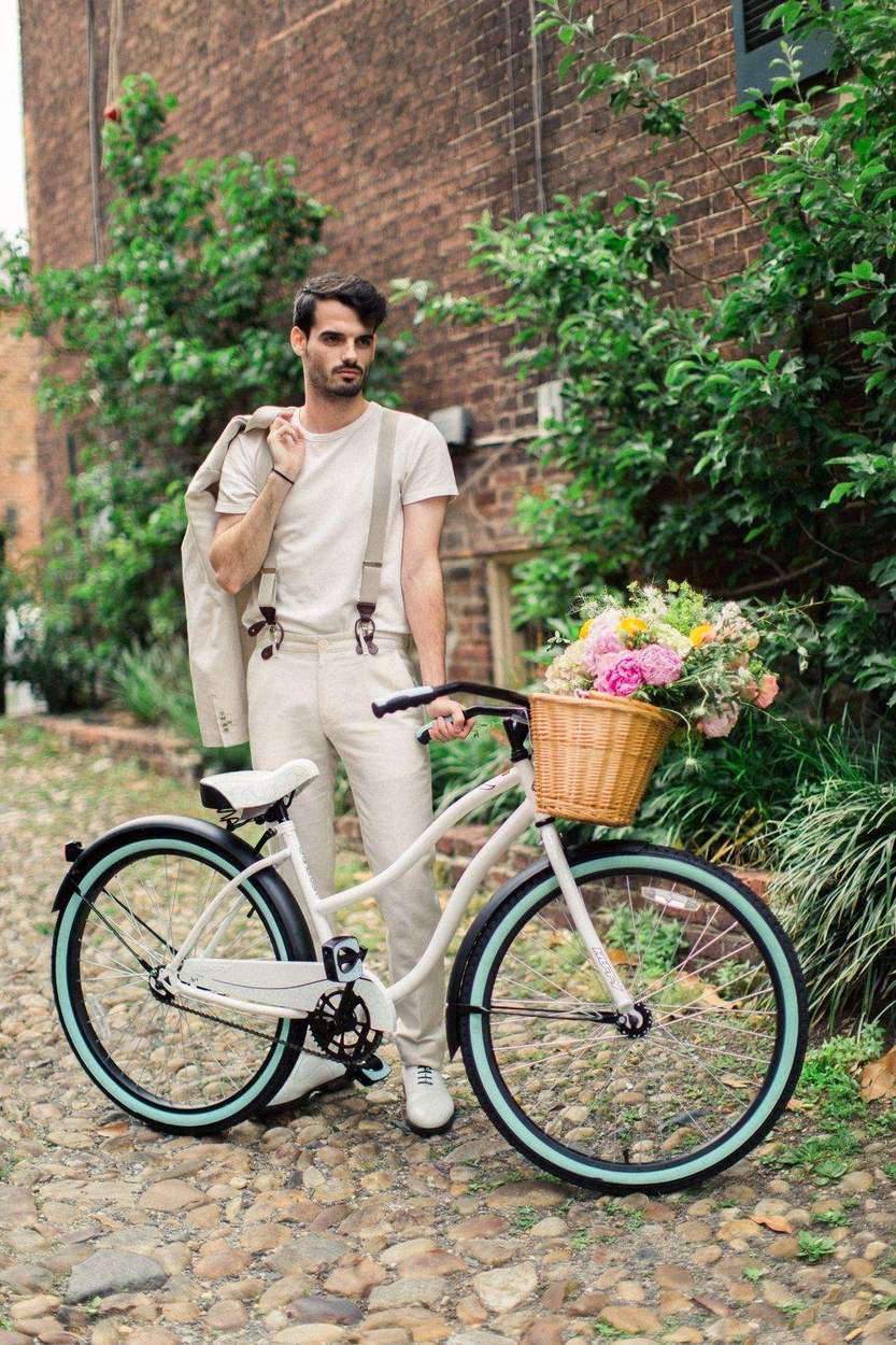Man in cream trousers and suspenders holding a suit jacket over his shoulder beside a white bicycle with mint-green tires and a flower basket.