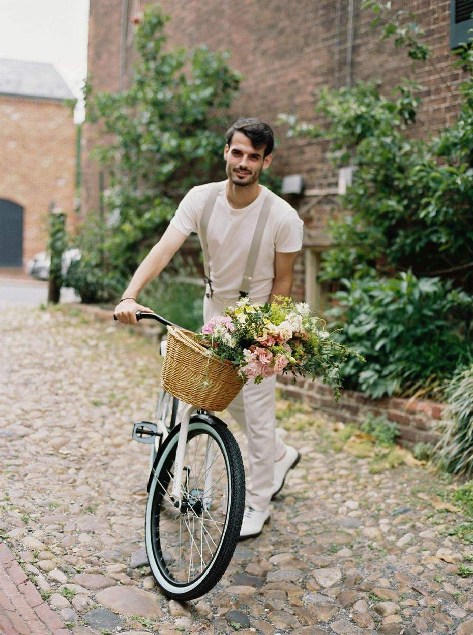 Smiling man in a cream T-shirt and suspenders walking a bicycle with a flower basket across cobblestones beside a brick wall.