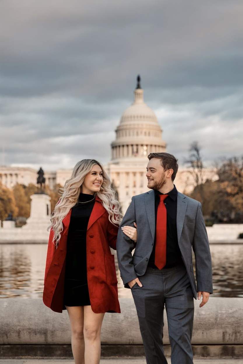 Couple walking arm in arm at the Capitol Reflecting Pool with the US Capitol dome rising behind them, she in a red coat and he in a gray suit.