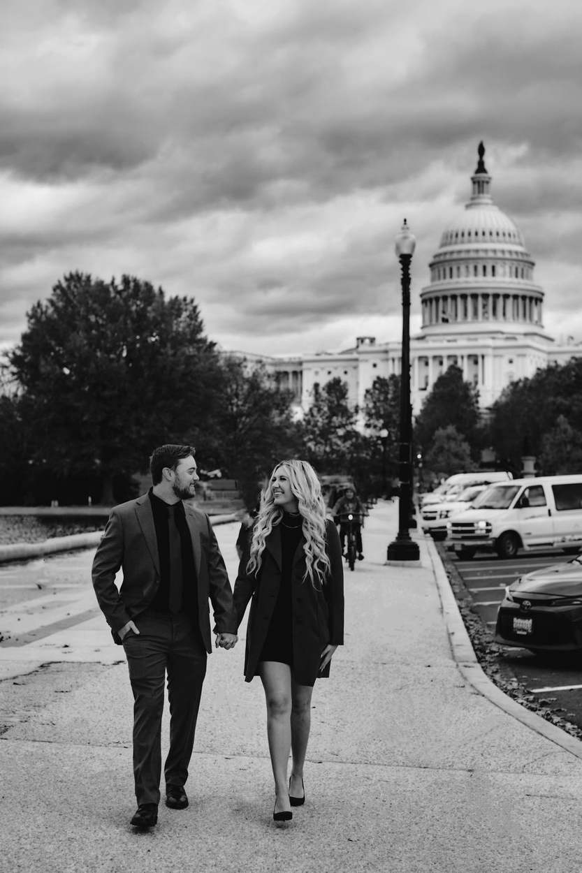 Black and white photo of a couple holding hands on a path with the US Capitol dome and a lamp post behind them under dramatic clouds.