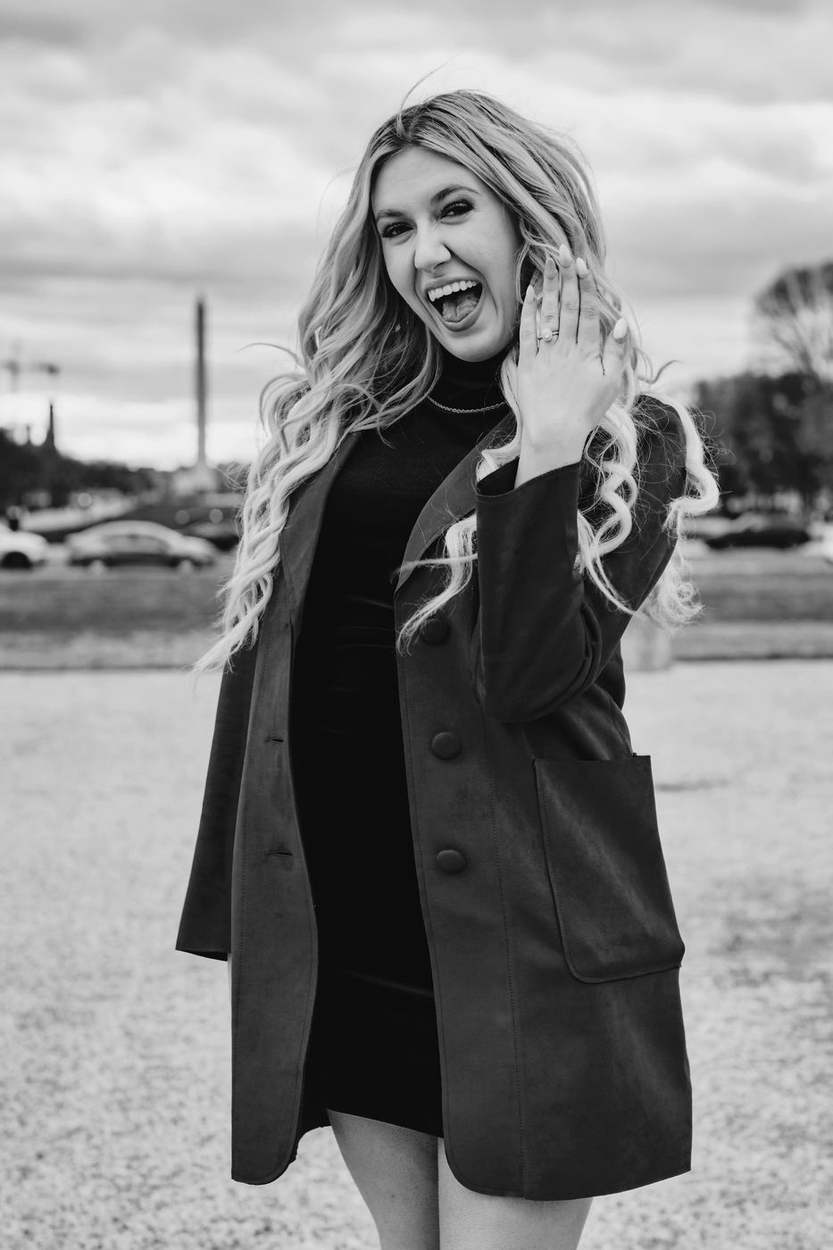 Black and white portrait of a woman laughing as she shows her engagement ring, standing near the Capitol Reflecting Pool.