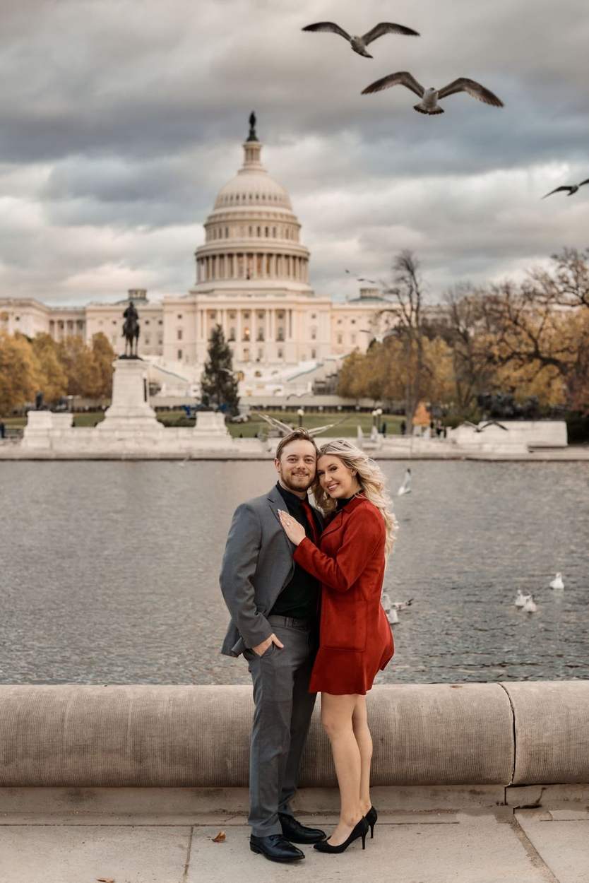 Couple embracing at the stone edge of the Capitol Reflecting Pool with the US Capitol dome and birds in flight behind them.