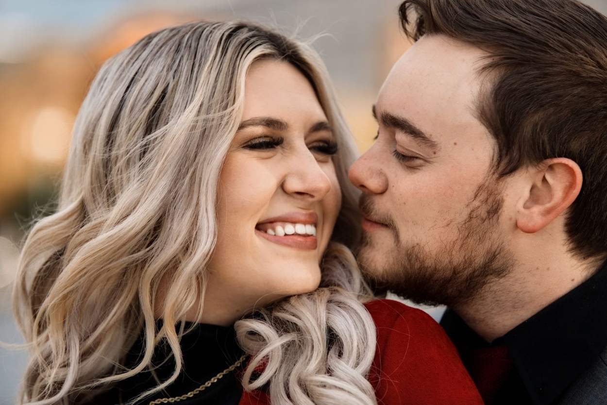 Close portrait of a smiling woman with wavy blonde hair as her fiancé kisses her cheek in warm autumn light.