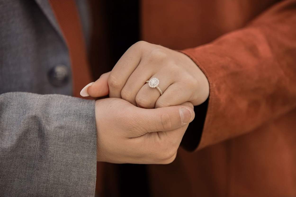 Detail of a couple's joined hands with a diamond engagement ring, her in a rust-red sleeve and him in a gray jacket.