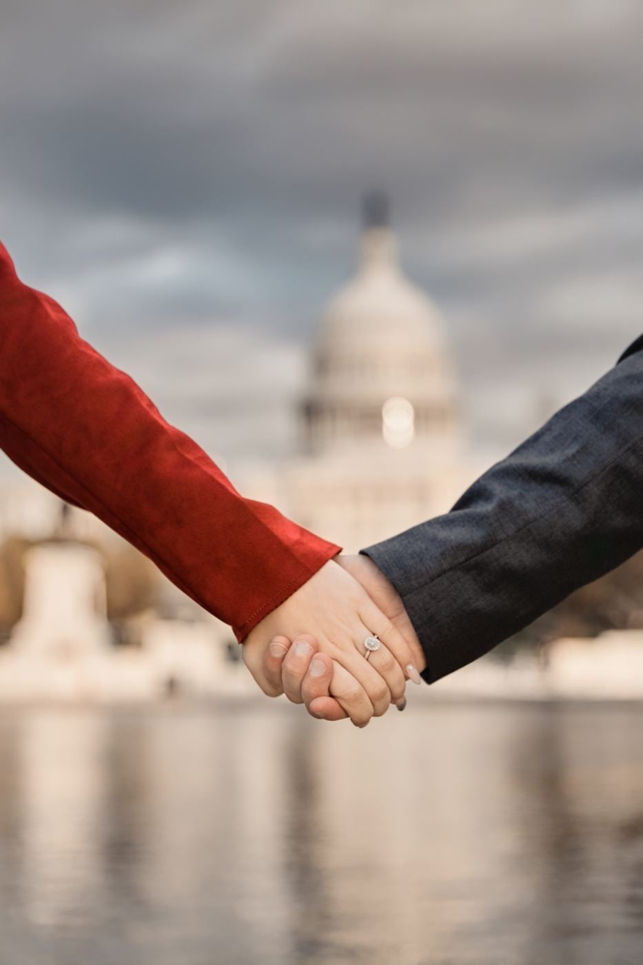 Close view of a couple's joined hands with an engagement ring, the US Capitol dome softly blurred behind them.