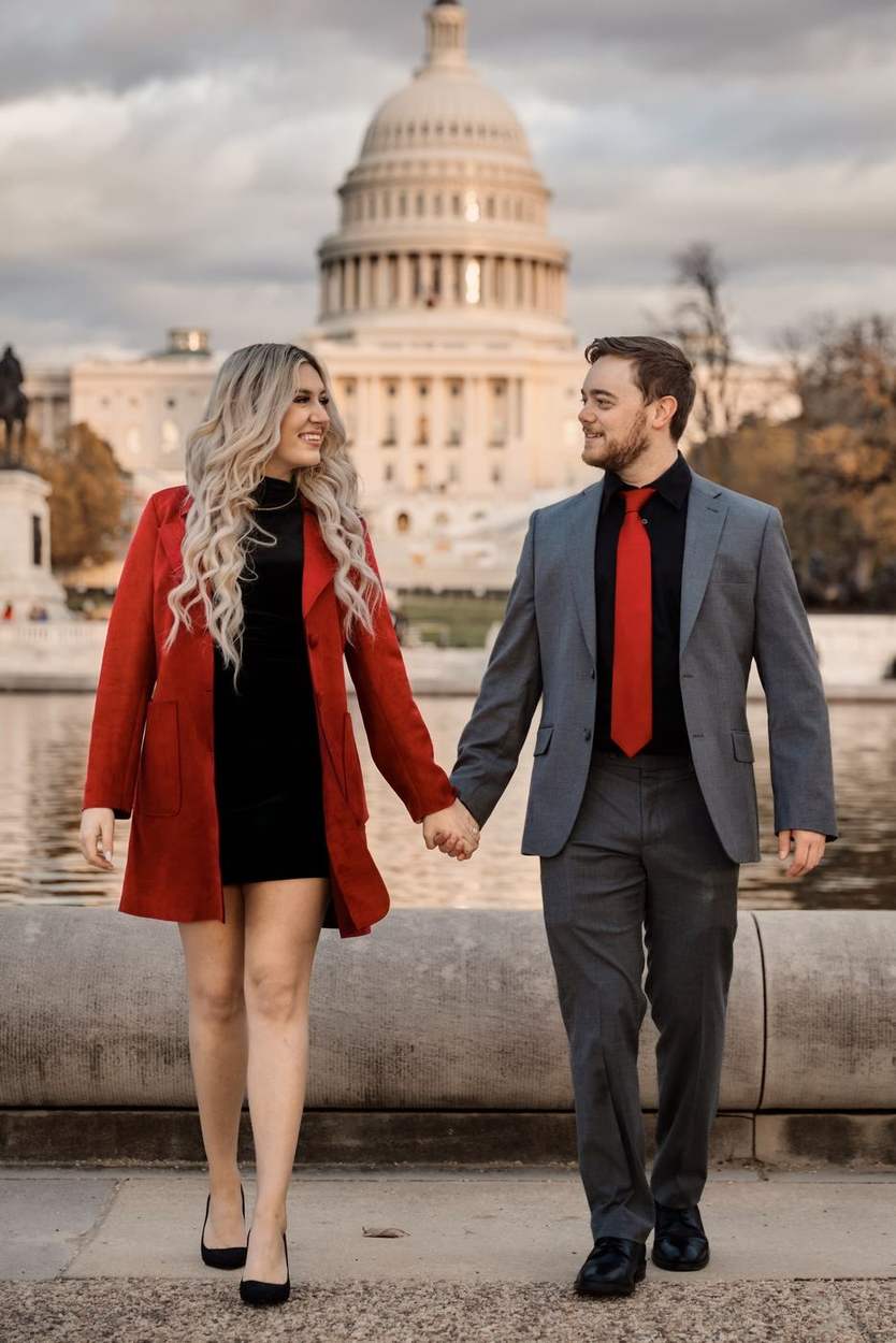 Couple holding hands and walking toward the camera along the stone edge of the Capitol Reflecting Pool with the US Capitol dome behind them.