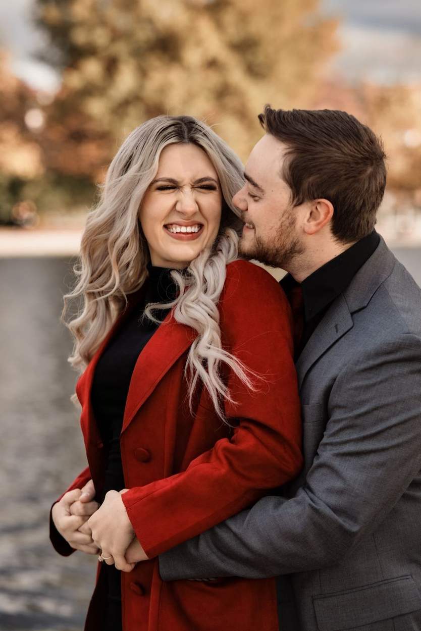 Laughing woman in a red coat held from behind by her fiancé in a gray suit beside the water, autumn leaves blurred behind them.