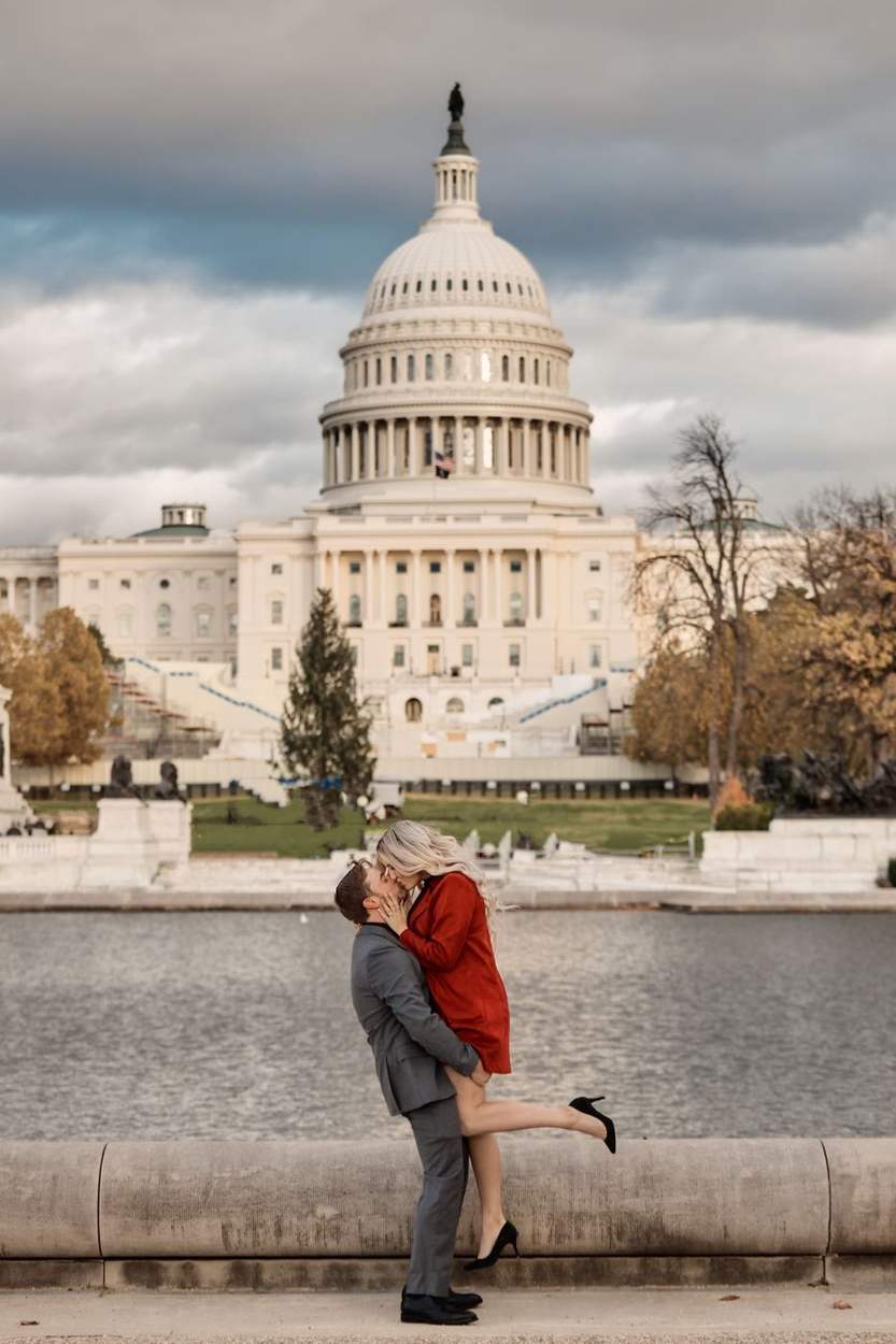 Man lifting his fiancée in a red coat for a kiss beside the Capitol Reflecting Pool, with the US Capitol dome and autumn trees behind them.