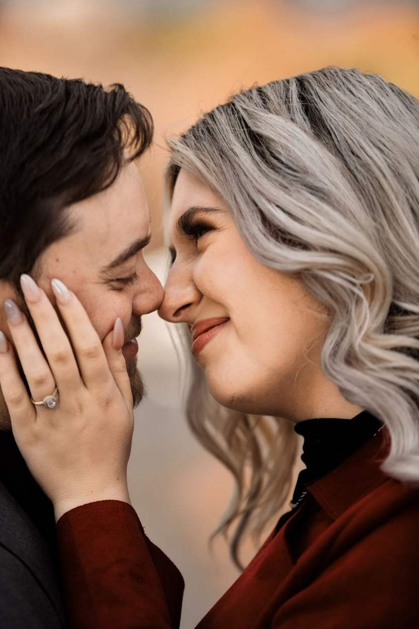 Couple nose to nose with her hand on his cheek and an engagement ring on show, warm autumn color glowing behind them.