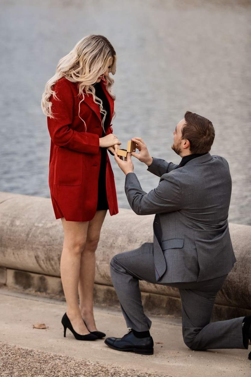 Man in a gray suit kneeling with an open ring box as a woman in a red coat looks down at him beside the Capitol Reflecting Pool.