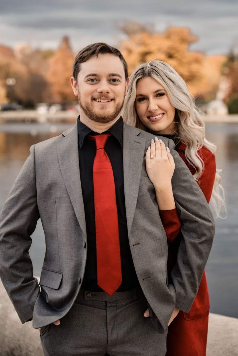 Smiling man in a gray suit and red tie with his fiancée in a red coat leaning on his shoulder, autumn trees and water behind them.