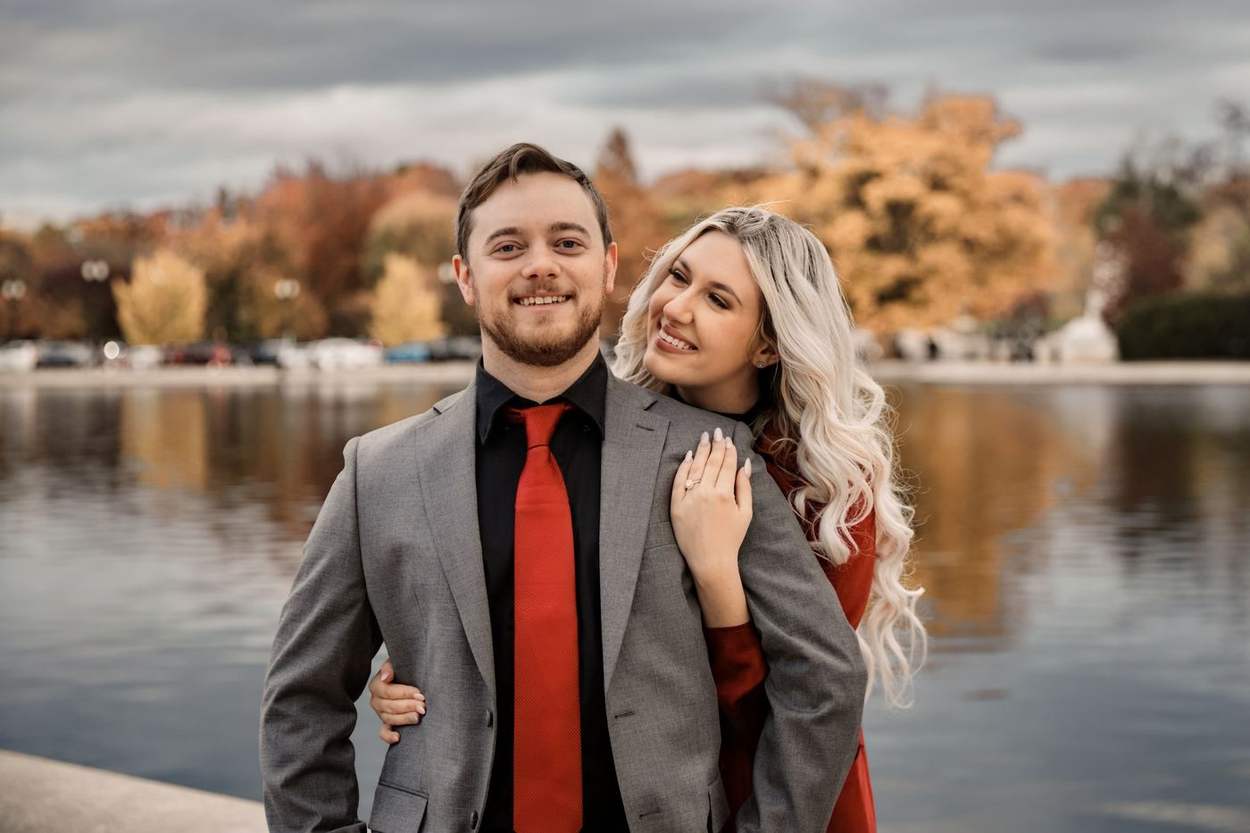 Smiling couple standing close beside the Capitol Reflecting Pool, he in a gray suit with a red tie and she in a red coat, autumn trees across the water.