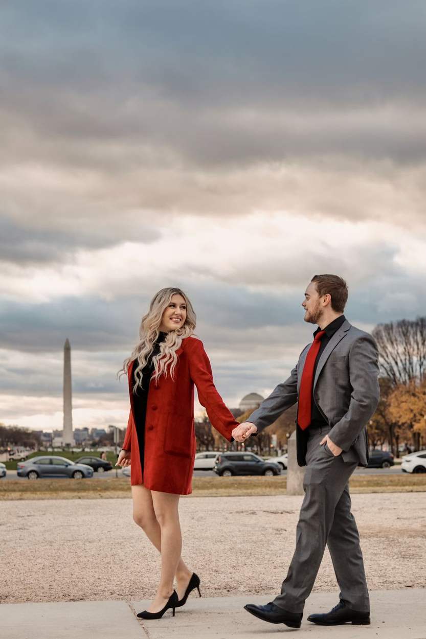 Couple holding hands and walking beside the Capitol Reflecting Pool with the Washington Monument in the distance under a cloudy sky.