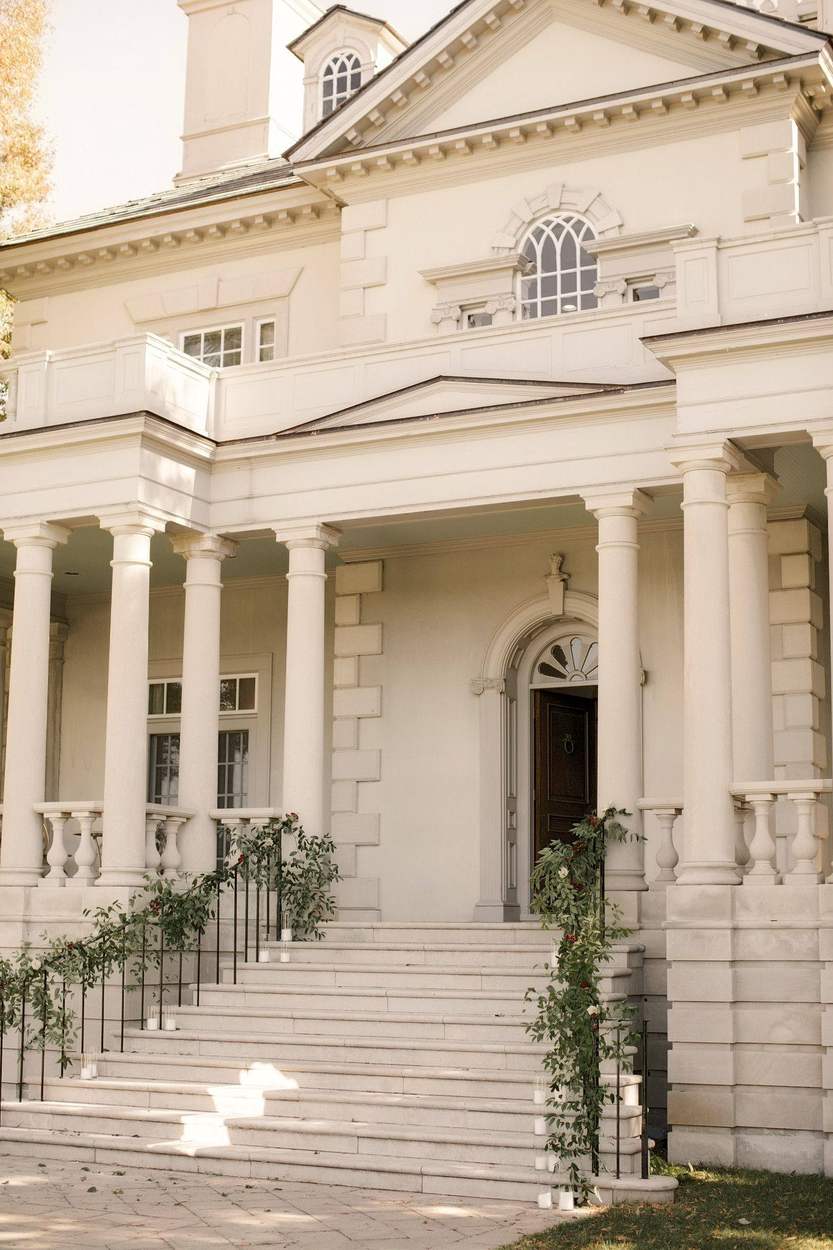 The cream-colored columned entrance of a grand estate, with an arched wooden door at the top of wide stone steps lined with greenery, red berries and pillar candles.
