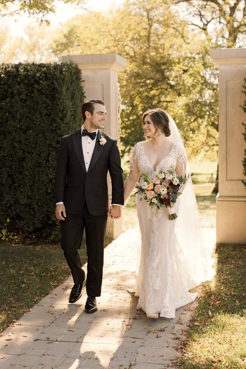 A smiling bride and groom in a black tuxedo walk hand in hand along a brick path between stone pillars and clipped hedges in warm autumn light, looking at each other.