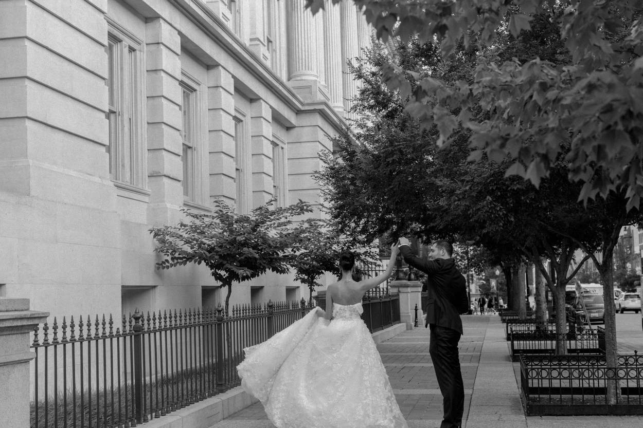 Black-and-white photograph of a groom twirling his bride in a tulle gown on a city sidewalk beside a stately stone building, iron railings and street trees.