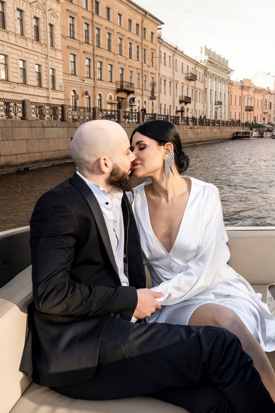 Groom in a black tuxedo jacket and bride in a white satin dress kissing on a boat, a canal embankment and pastel buildings behind them