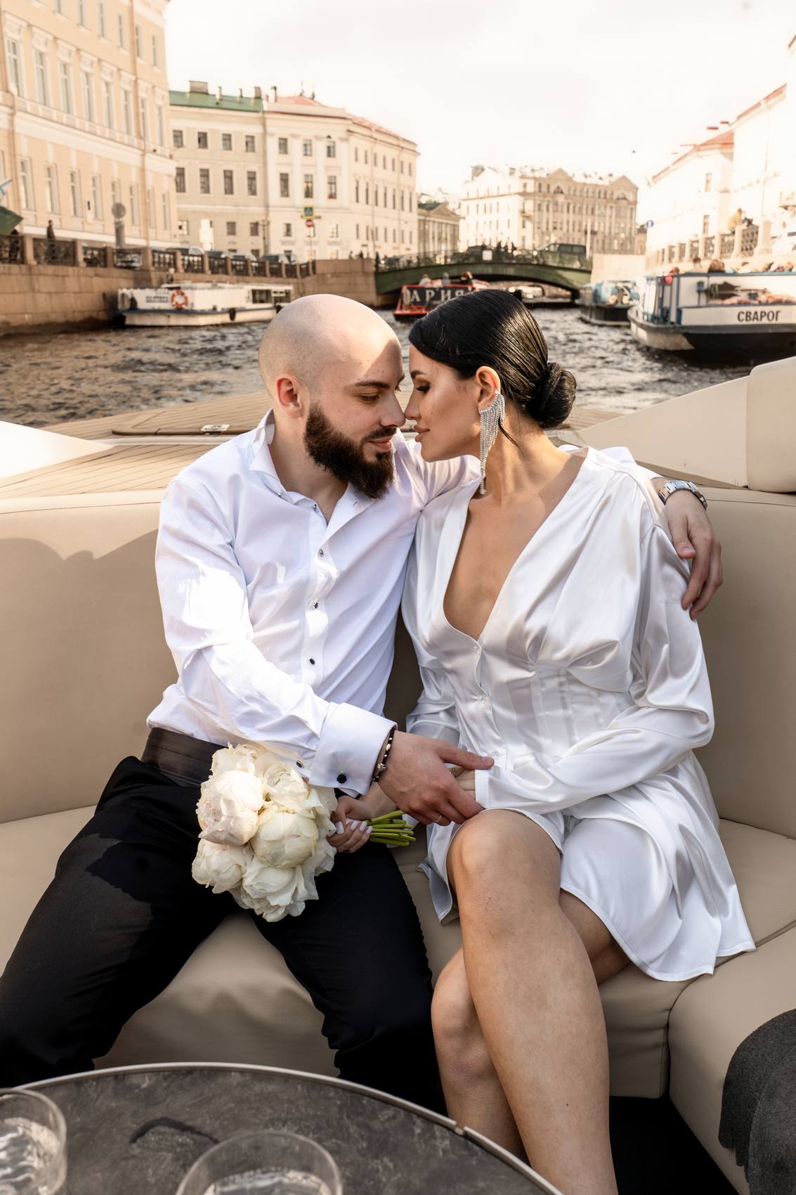 Couple nose to nose on a boat seat holding white peonies, a canal lined with historic buildings and a green bridge behind them