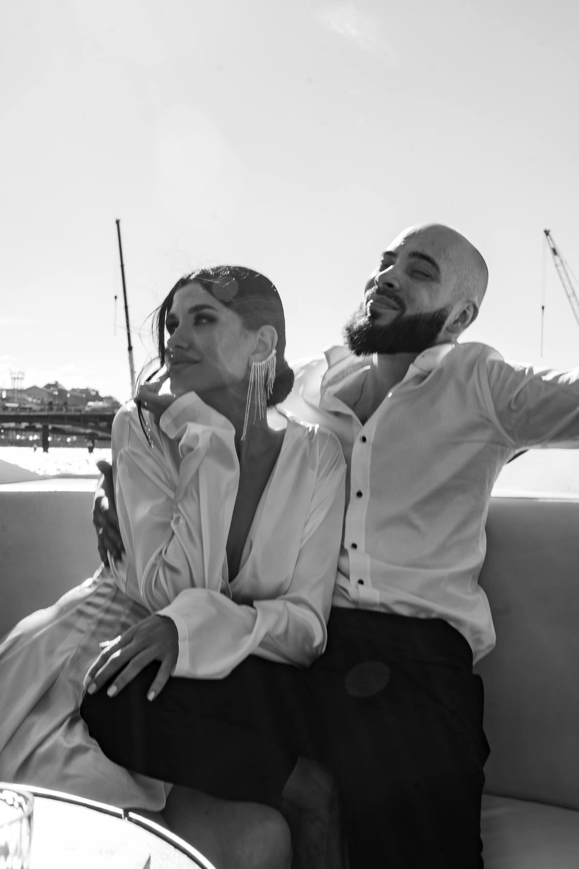 Black and white photograph of a couple on a boat in bright light, she looking away with her chin on her hand and he leaning back with his arm stretched out, cranes behind them
