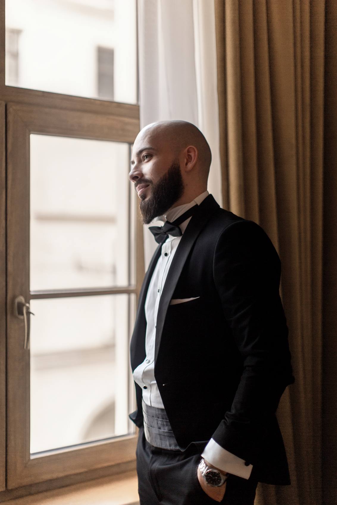 Groom in a black tuxedo with a bow tie and cummerbund looking out of a tall window with his hands in his pockets, a brown curtain beside him