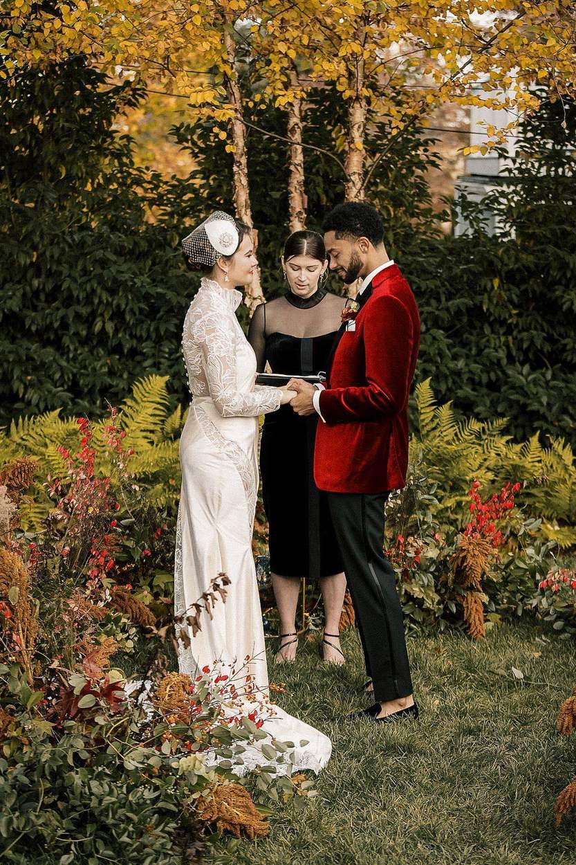 A bride in a high-neck lace gown and a groom in a red velvet jacket hold hands as an officiant reads to them in a garden of ferns and autumn foliage.