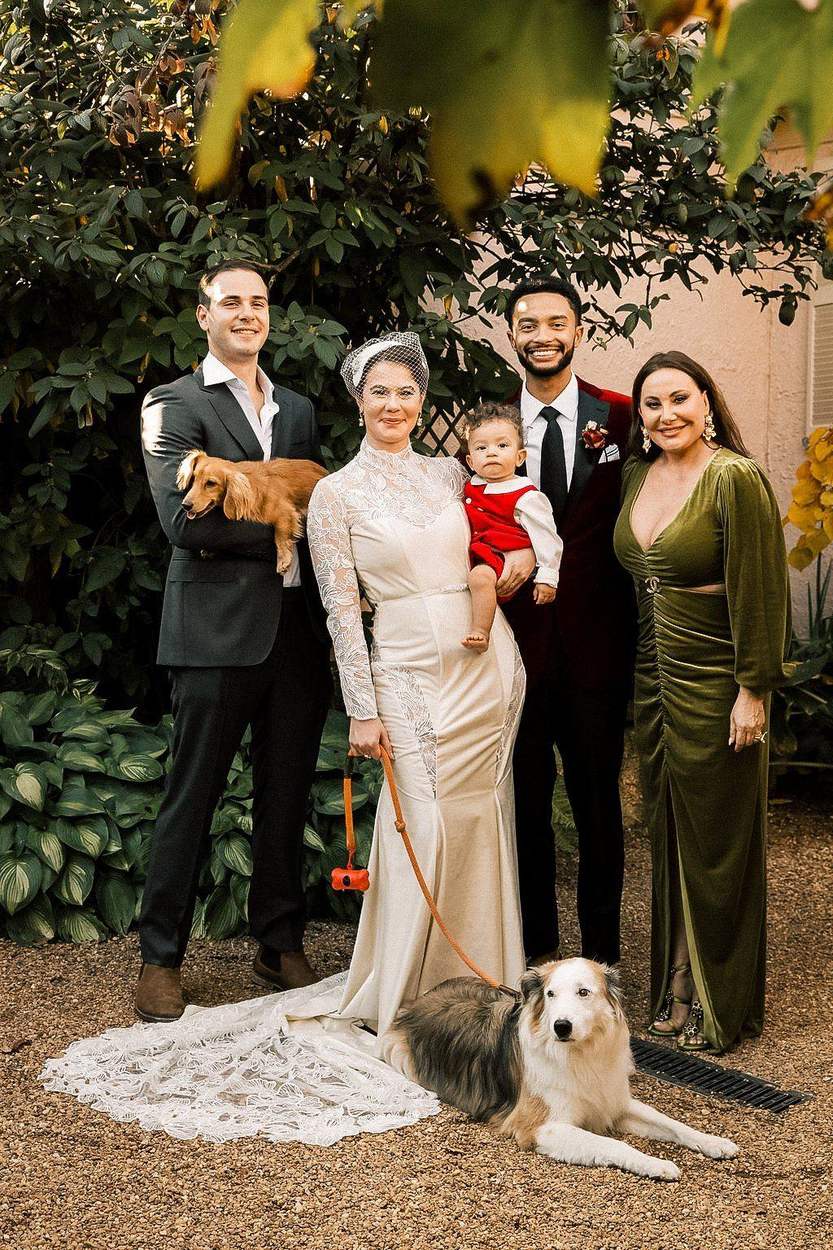 A bride and groom with their baby, two relatives and two dogs pose together in a garden courtyard in autumn.