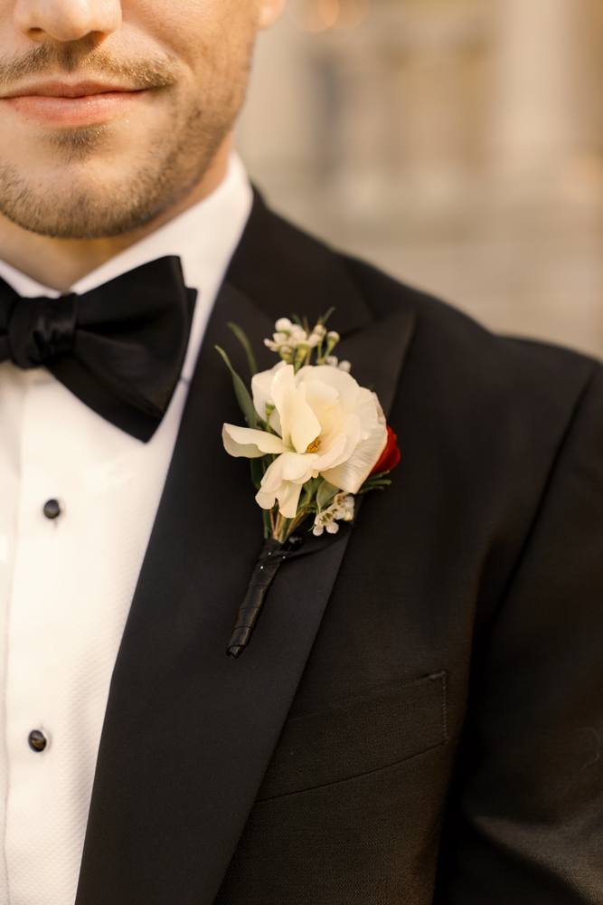 Close view of a white rose boutonniere pinned to a black tuxedo lapel beside a black bow tie