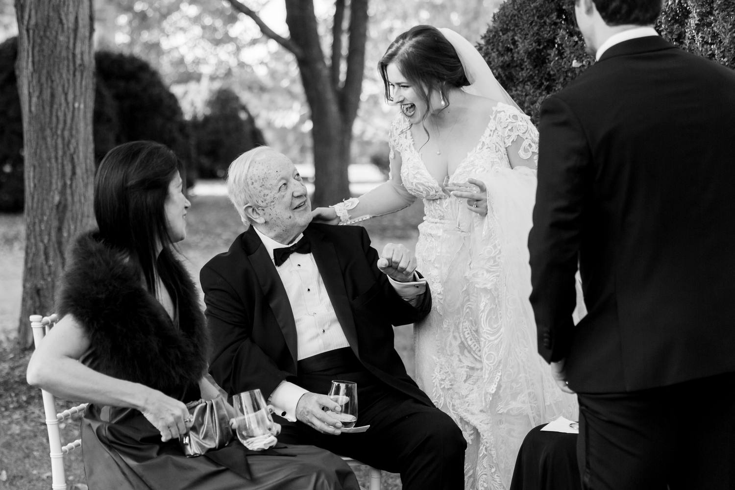 Black and white photo of the bride laughing as she leans in to greet a seated older guest in a tuxedo