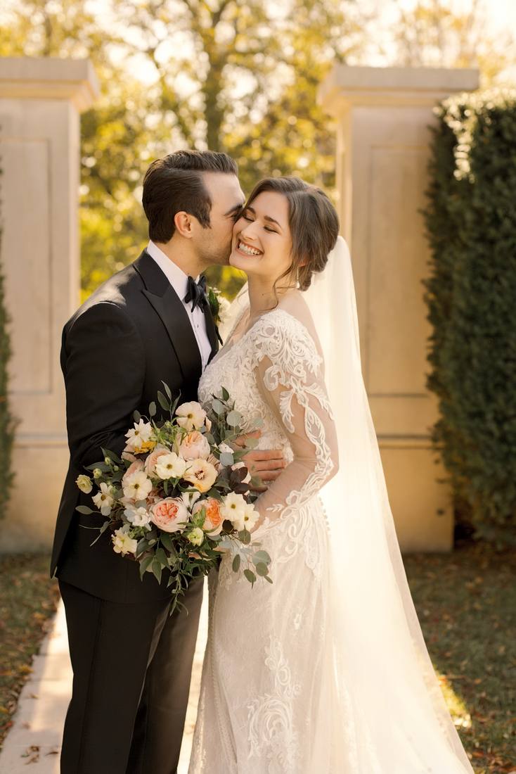 Bride and groom embracing in golden light beside a stone pillar and a tall clipped hedge