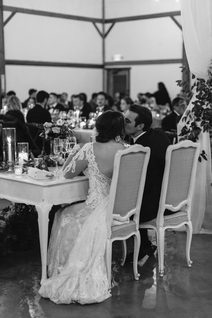 Black and white photo of the couple kissing at their sweetheart table, seen from behind, guests seated at candlelit tables beyond